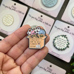 May include: Enamel pin showing a box overflowing with pink, blue, and white flowers. The pin reads "Consider How The Wild Flowers Grow" with a heart and the word "Luke" below. The pin is on a pink card.