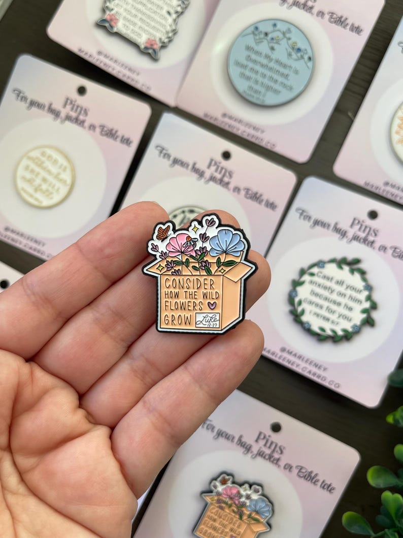 May include: Enamel pin showing a box overflowing with pink, blue, and white flowers. The pin reads "Consider How The Wild Flowers Grow" with a heart and the word "Luke" below. The pin is on a pink card.