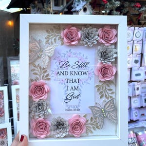 May include: A white framed shadow box with a floral design. The box features pink and gray paper roses, glittery butterflies, and the text "Be Still and Know That I Am God" from Psalm 46:10.