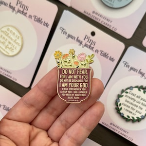May include: A gold-coloured pin featuring a floral design and the text "DO NOT FEAR, FOR I AM WITH YOU. I AM YOUR GOD." The pin is held in a hand, with other pins visible in the background.