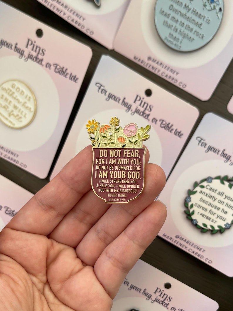 May include: A gold-coloured pin featuring a floral design and the text "DO NOT FEAR, FOR I AM WITH YOU. I AM YOUR GOD." The pin is held in a hand, with other pins visible in the background.