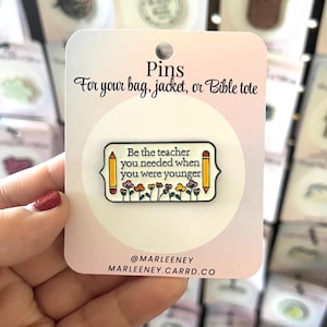 May include: A rectangular enamel pin with the text "Be the teacher you needed when you were younger" and colorful flowers. The pin is on a pink card with the word "Pins" and the phrase "For your bag, jacket, or Bible tote."
