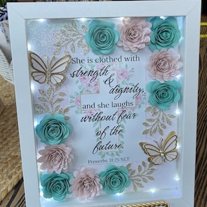 May include: White framed art piece with the text "She is clothed with strength & dignity, and she laughs without fear of the future. Proverbs 31:25 NLT." Features paper flowers in teal and pink, gold butterflies, and fairy lights.