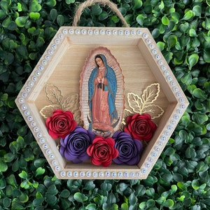 Puede incluir: Un colgante de pared hexagonal de madera con una imagen de Nuestra Se&ntilde;ora de Guadalupe. El marco est&aacute; adornado con pedrer&iacute;a y flores de papel en rojo y morado. Acentos de hojas doradas y una cuerda de yute para colgar completan el dise&ntilde;o.