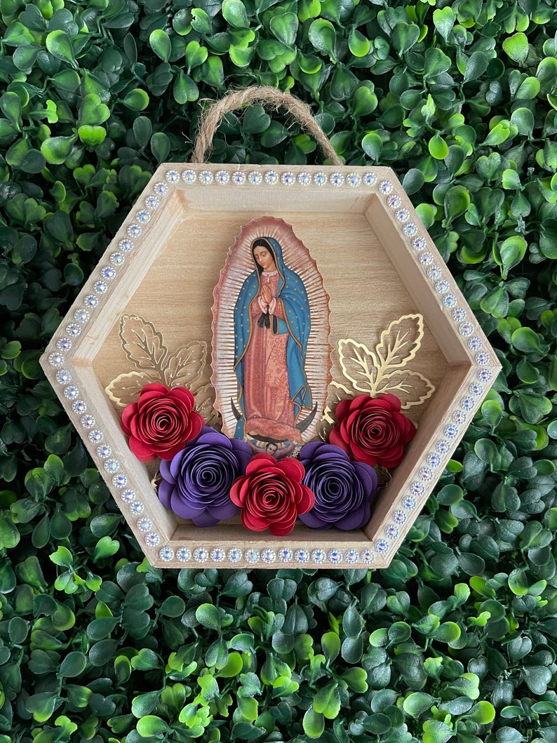 Puede incluir: Un colgante de pared hexagonal de madera con una imagen de Nuestra Se&ntilde;ora de Guadalupe. El marco est&aacute; adornado con pedrer&iacute;a y flores de papel en rojo y morado. Acentos de hojas doradas y una cuerda de yute para colgar completan el dise&ntilde;o.