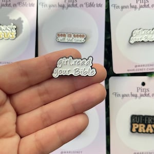 May include: A hand holds a white and silver enamel pin that says "girl read your Bible" in a cursive font. Other pins in the background have phrases like "God is good" and "But first, pray". The pins are on pink cards.