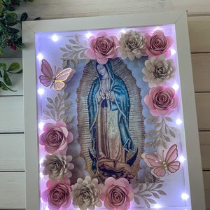 May include: A white frame with a light inside, featuring a religious image of the Virgin Mary surrounded by pink and beige paper flowers and butterflies. The frame is decorated with glitter and leaves.