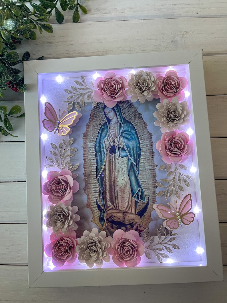 May include: A white frame with a light inside, featuring a religious image of the Virgin Mary surrounded by pink and beige paper flowers and butterflies. The frame is decorated with glitter and leaves.