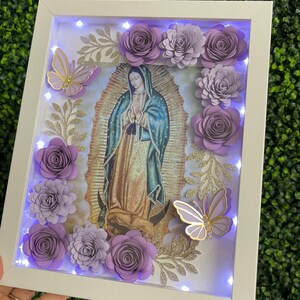 May include: A white frame with a light inside, featuring a religious image of the Virgin Mary with purple paper flowers and butterflies around the image.