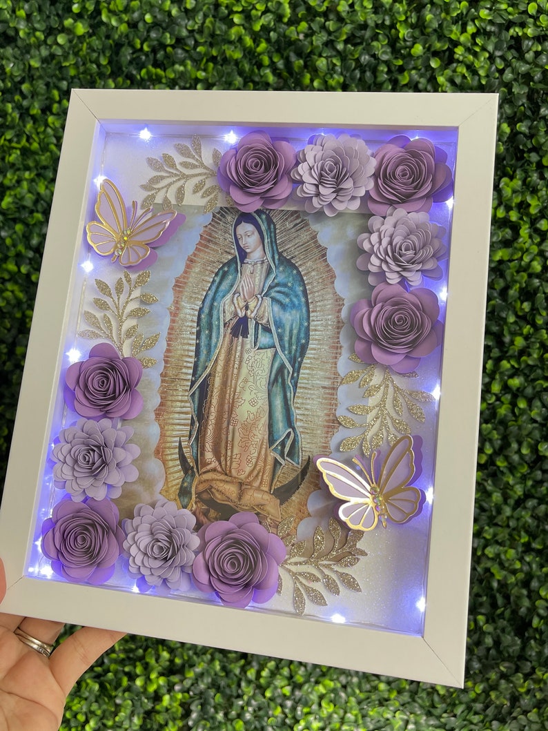 May include: A white frame with a light inside, featuring a religious image of the Virgin Mary with purple paper flowers and butterflies around the image.