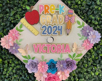 May include: A white graduation cap decorated with colorful paper flowers, a red apple, a pencil, a ruler, and the text "Pre-K Grad 2024 Victoria".