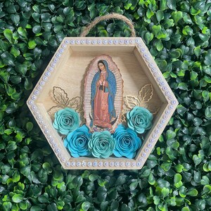 Puede incluir: Un colgante de pared hexagonal de madera con una imagen de Nuestra Se&ntilde;ora de Guadalupe. El marco est&aacute; adornado con pedrer&iacute;a y detalles de hojas doradas. Rosas de papel turquesa est&aacute;n dispuestas en la parte inferior. Una cuerda de yute est&aacute; unida para colgar.