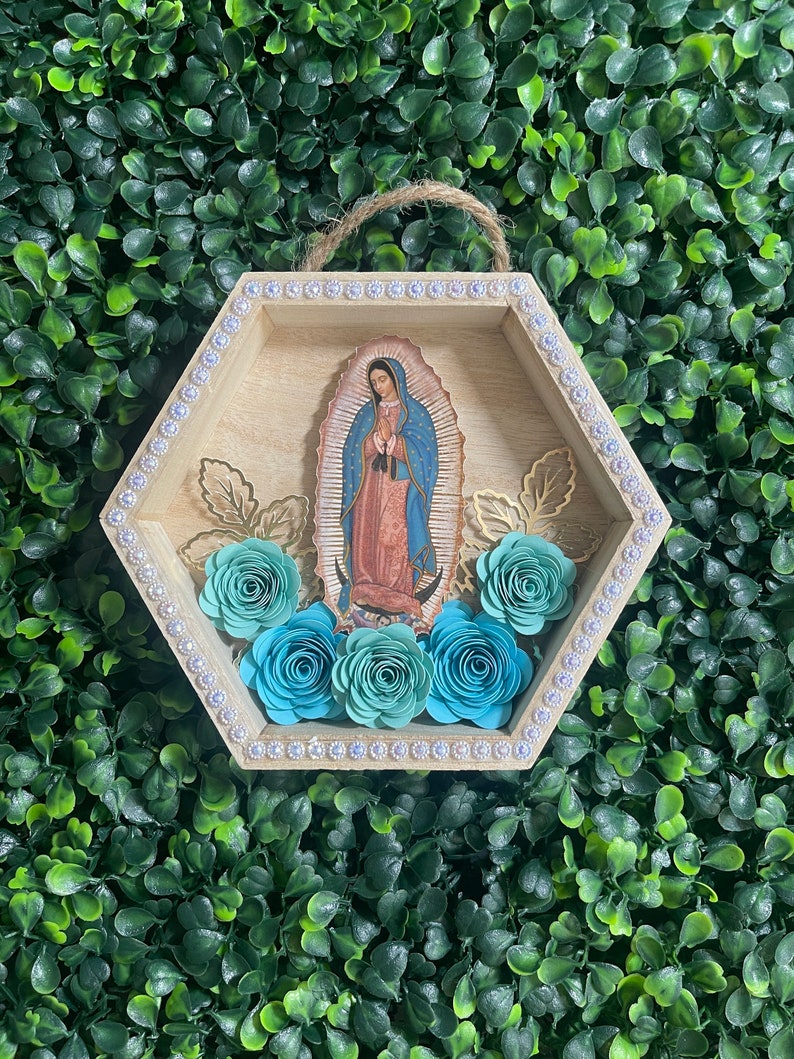 Puede incluir: Un colgante de pared hexagonal de madera con una imagen de Nuestra Se&ntilde;ora de Guadalupe. El marco est&aacute; adornado con pedrer&iacute;a y detalles de hojas doradas. Rosas de papel turquesa est&aacute;n dispuestas en la parte inferior. Una cuerda de yute est&aacute; unida para colgar.