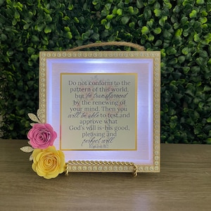 May include: A decorative illuminated frame with a Bible verse. The frame has a gold border with a string handle. It features a quote from Romans 12:2 and is adorned with pink and yellow paper flowers.