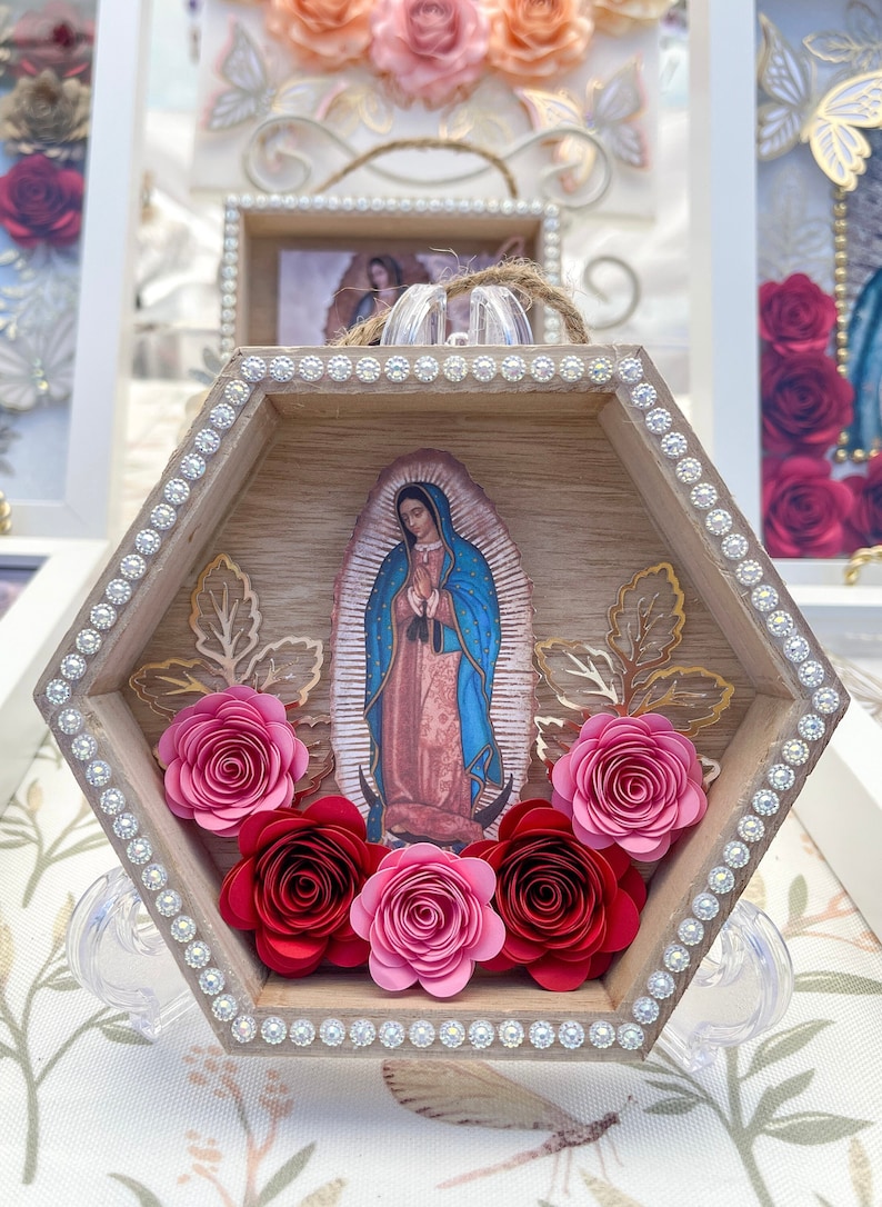 Puede incluir: Una caja de sombra hexagonal de madera con una imagen de Nuestra Se&ntilde;ora de Guadalupe. El marco est&aacute; decorado con pedrer&iacute;a y rosas de papel en tonos rojos y rosas. Acentos de pan de oro y un soporte transparente completan el dise&ntilde;o.