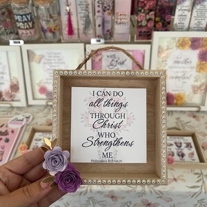 May include: A square wooden framed sign with the text "I CAN DO all things THROUGH Christ WHO Strengthens ME" in black script. The frame is decorated with rhinestones and paper flowers. Other decorative items are visible in the background.
