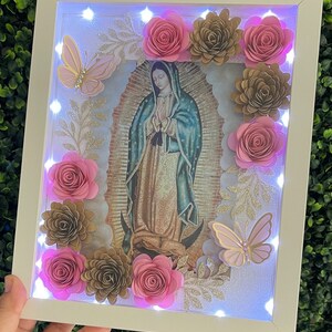 May include: A white frame with a light-up image of the Virgin Mary surrounded by pink and gold paper flowers and butterflies.