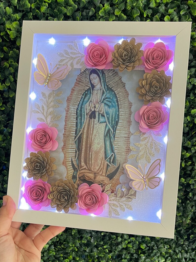 May include: A white frame with a light-up image of the Virgin Mary surrounded by pink and gold paper flowers and butterflies.