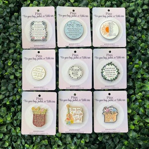 May include: Nine enamel pins on cards with inspirational quotes and floral designs. The pins are in various shapes, including circles and rectangles. The text on the cards reads "Pins for your bag, jacket, or Bible tote."
