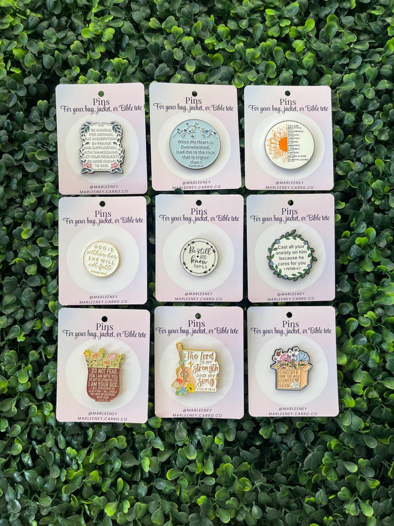 May include: Nine enamel pins on cards with inspirational quotes and floral designs. The pins are in various shapes, including circles and rectangles. The text on the cards reads "Pins for your bag, jacket, or Bible tote."