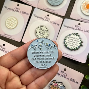 May include: A selection of enamel pins with motivational quotes and floral motifs. The pins are circular and display phrases such as "When My Heart is Overwhelmed" and "Be Still and Know." They are presented on small cards.