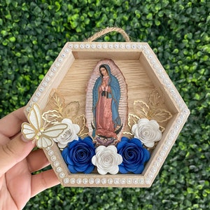 Puede incluir: Un colgante de pared hexagonal de madera con una imagen de Nuestra Se&ntilde;ora de Guadalupe. El marco est&aacute; decorado con pedrer&iacute;a, rosas de papel en blanco y azul, hojas doradas y una mariposa brillante. Se adjunta una cuerda de yute para colgar.