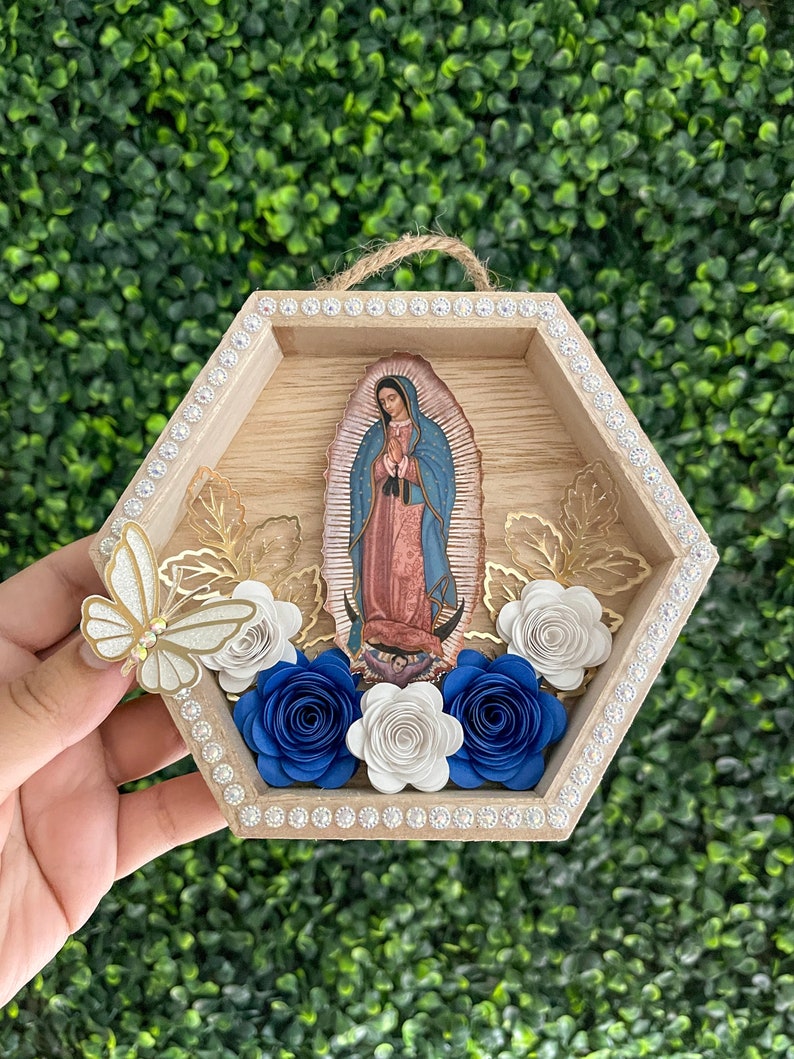 Puede incluir: Un colgante de pared hexagonal de madera con una imagen de Nuestra Se&ntilde;ora de Guadalupe. El marco est&aacute; decorado con pedrer&iacute;a, rosas de papel en blanco y azul, hojas doradas y una mariposa brillante. Se adjunta una cuerda de yute para colgar.