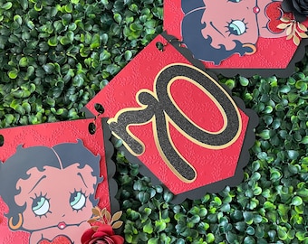 May include: Red and black birthday banner with the number 70 in gold glitter, featuring a cartoon character with red lips and black hair. The banner is decorated with black and red paper flowers.