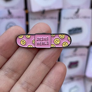May include: A pink enamel pin in the shape of a bandage. The pin features yellow smiley faces and the words "JESUS HEALS" in white. The pin has a black border.