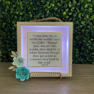 May include: A framed sign with a Bible verse, featuring a light-up frame and decorative paper flowers in shades of blue and white. The text reads, "Come now, let us settle the matter..." from Isaiah 1:18.