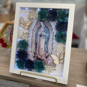 May include: A white frame with a light-up image of the Virgin Mary surrounded by teal and navy blue paper flowers, gold glitter, and gold butterflies.