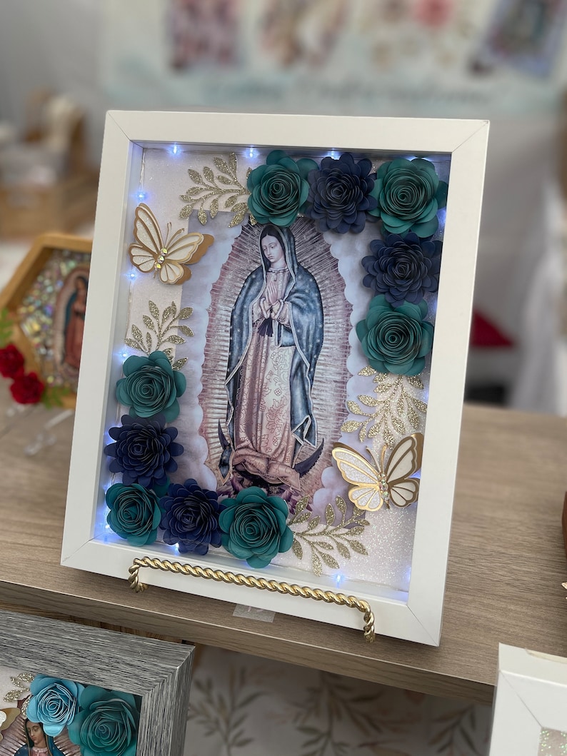 May include: A white frame with a light-up image of the Virgin Mary surrounded by teal and navy blue paper flowers, gold glitter, and gold butterflies.