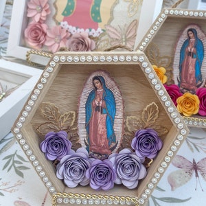 Puede incluir: Arte de pared hexagonal de madera con una imagen de Nuestra Se&ntilde;ora de Guadalupe. El marco est&aacute; adornado con pedrer&iacute;a y flores de papel en tonos morados. Los detalles dorados y una cuerda para colgar completan el dise&ntilde;o. Otras obras de arte enmarcadas similares son visibles en el fondo.