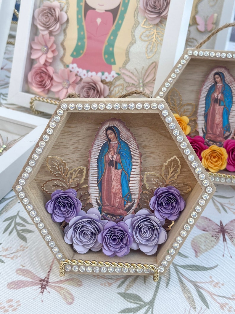 Puede incluir: Arte de pared hexagonal de madera con una imagen de Nuestra Se&ntilde;ora de Guadalupe. El marco est&aacute; adornado con pedrer&iacute;a y flores de papel en tonos morados. Los detalles dorados y una cuerda para colgar completan el dise&ntilde;o. Otras obras de arte enmarcadas similares son visibles en el fondo.