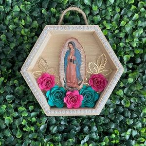 Puede incluir: Un colgante de pared hexagonal de madera con una imagen de Nuestra Se&ntilde;ora de Guadalupe. El marco est&aacute; adornado con pedrer&iacute;a y detalles de hojas doradas. Rosas de papel rosas y turquesas est&aacute;n dispuestas en la parte inferior, con una cuerda de yute para colgar.