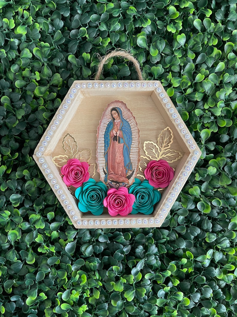 Puede incluir: Un colgante de pared hexagonal de madera con una imagen de Nuestra Se&ntilde;ora de Guadalupe. El marco est&aacute; adornado con pedrer&iacute;a y detalles de hojas doradas. Rosas de papel rosas y turquesas est&aacute;n dispuestas en la parte inferior, con una cuerda de yute para colgar.