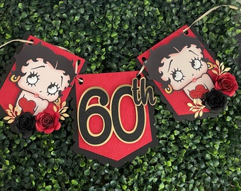 May include: Red and black birthday banner with a cartoon character and the number 60 with the word "th" next to it. The banner is decorated with black and red flowers.