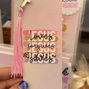 May include: A pink bookmark with the words "Jesus loves, forgives, saves" in black script. It has a pink tassel and a gold and silver cross charm. The bookmark is held by a hand with blue and silver nail polish.