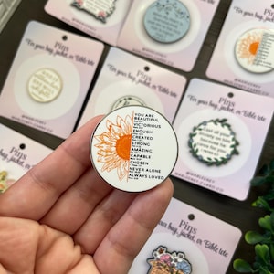 May include: A selection of enamel pins displayed on pink cards. One pin has a sunflower design and the words "YOU ARE BEAUTIFUL, VICTORIOUS, ENOUGH, CREATED, STRONG, AMAZING, CAPABLE, CHOSEN, NEVER ALONE, ALWAYS LOVED."