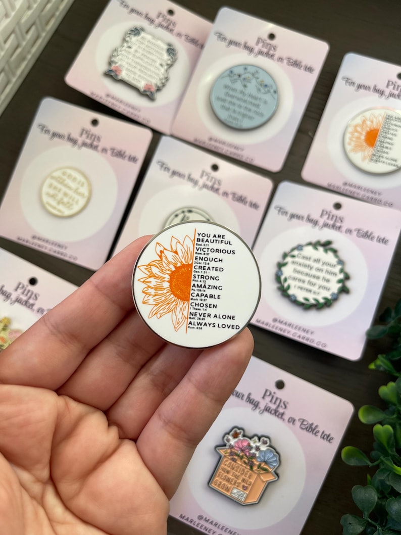 May include: A selection of enamel pins displayed on pink cards. One pin has a sunflower design and the words "YOU ARE BEAUTIFUL, VICTORIOUS, ENOUGH, CREATED, STRONG, AMAZING, CAPABLE, CHOSEN, NEVER ALONE, ALWAYS LOVED."