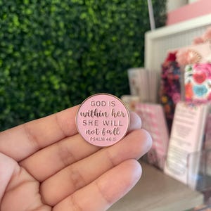 May include: A round, light pink enamel pin with a gold-toned metal border. The pin features the embossed text "GOD IS within her SHE WILL not fall PSALM 46:5" in a combination of block and script lettering, centered on the pink surface.