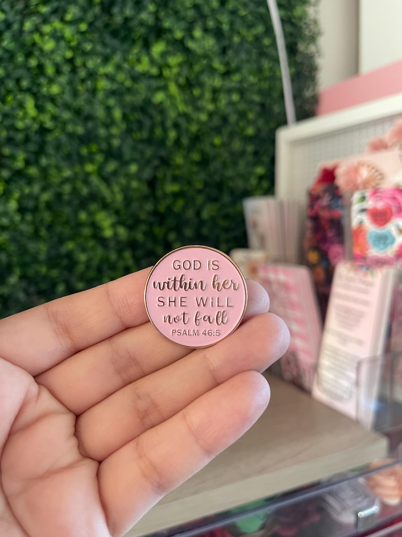 May include: A round, light pink enamel pin with a gold-toned metal border. The pin features the embossed text "GOD IS within her SHE WILL not fall PSALM 46:5" in a combination of block and script lettering, centered on the pink surface.