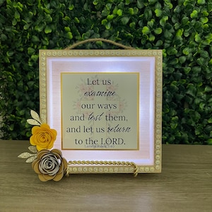 May include: A framed inspirational sign with the text "Let us examine our ways and test them, and let us return to the LORD. Lamentations 3:40." The frame has a gold border, with yellow and brown paper flowers.