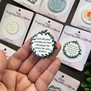 May include: A collection of enamel pins with motivational quotes and floral motifs. The pins display phrases such as "Cast all your anxiety on him" and "God is within her, she will not fall." Each pin is presented on a card labelled "Pins for your bag, jacket, or Bible tote."