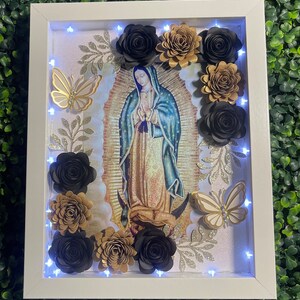 May include: A white framed picture with a religious image of the Virgin Mary surrounded by black and gold paper flowers and butterflies. The frame has a light string of white LED lights around the inside edge.