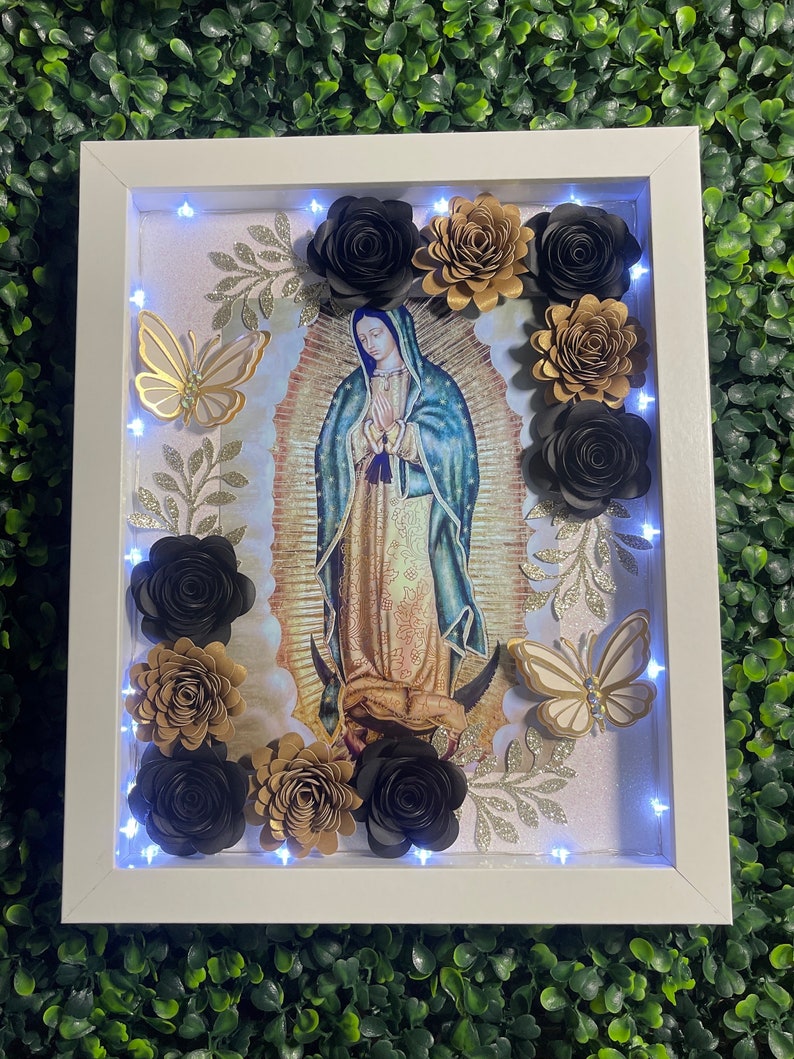 May include: A white framed picture with a religious image of the Virgin Mary surrounded by black and gold paper flowers and butterflies. The frame has a light string of white LED lights around the inside edge.
