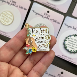 May include: A gold-coloured pin with the text "The Lord is my strength and my song" and a guitar illustration. The pin features colourful floral accents and is designed to be attached to a bag, jacket, or Bible tote.