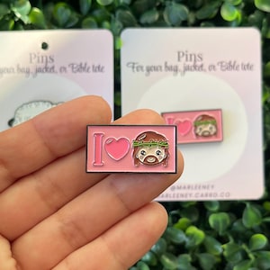 May include: Pink enamel pin with the text "I ❤️" and a cartoon image of Jesus wearing a crown of thorns. The pin is rectangular with a black border. The background includes a green leafy plant and a card that says "Pins for your bag, jacket, or Bible tote."