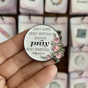 May include: A round enamel pin with the text "DON'T WORRY ABOUT ANYTHING; INSTEAD PRAY ABOUT EVERYTHING" and the Bible verse Philippians 4:6. The pin features pink floral accents and a black border.