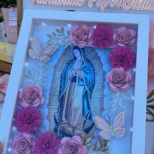 May include: A white frame with a light blue image of the Virgin Mary surrounded by pink paper flowers and butterflies. The frame has a sign above it that reads "Marleene Handmade Paper Creations".